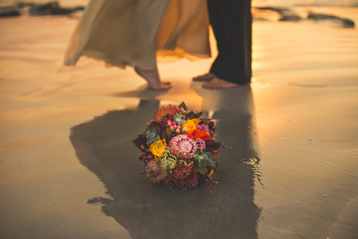 Bouquet de fleurs sur la plage et pieds du couple marié dans le fond. Photographe de Mariage Toulouse.